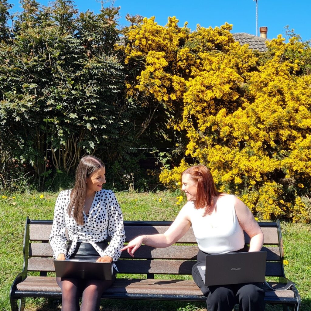 Youbee Emma and Youbee Becky looking at laptops on bench